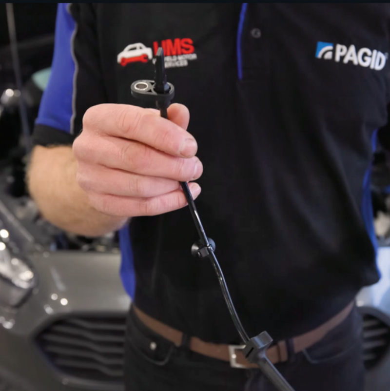 A close-up of a mechanic holding a black ABS wheel speed sensor cable, wearing a black and blue Pagid shirt with a UMS logo, standing in front of a grey car.