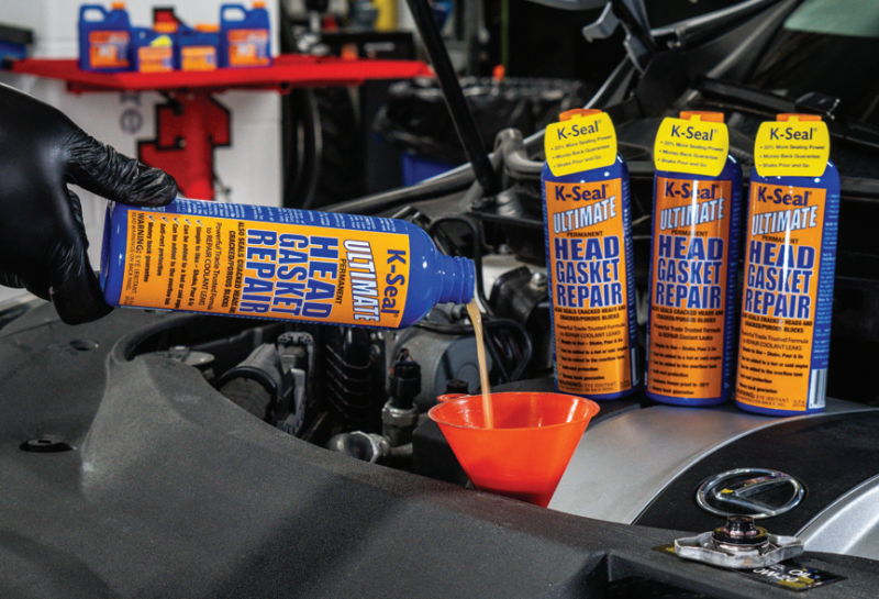 A mechanic wearing a black glove pouring K-Seal Ultimate Head Gasket Repair into a car engine using an orange funnel.