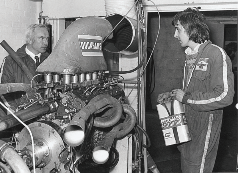 A vintage black and white photograph of two mechanics working on a racing engine featuring a Duckhams sponsor logo.