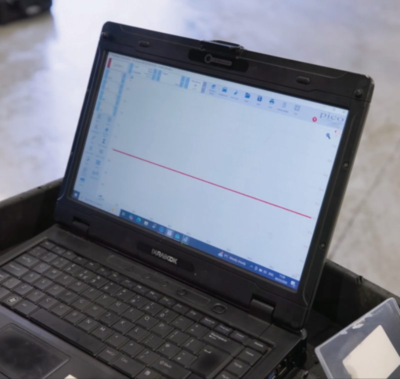 A rugged Durabook laptop inside an automotive garage running PicoScope software, showing a flat, straight red line on the screen indicating a lack of waveform signal from a faulty wheel speed sensor.