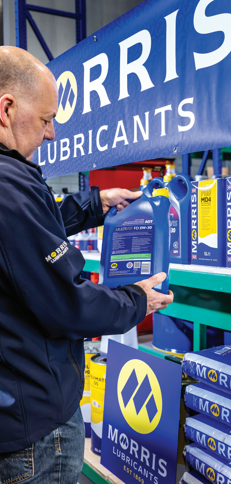 A man in a Morris Lubricants jacket selecting a large blue 5-litre jug of Multivis FD 5W-30 engine oil from a warehouse retail display shelf.