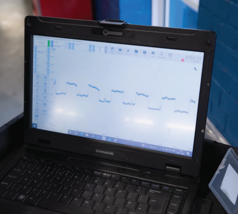 A rugged Durabook diagnostic laptop running PicoScope software inside a garage, displaying a clean digital square waveform from a properly functioning active wheel speed sensor.