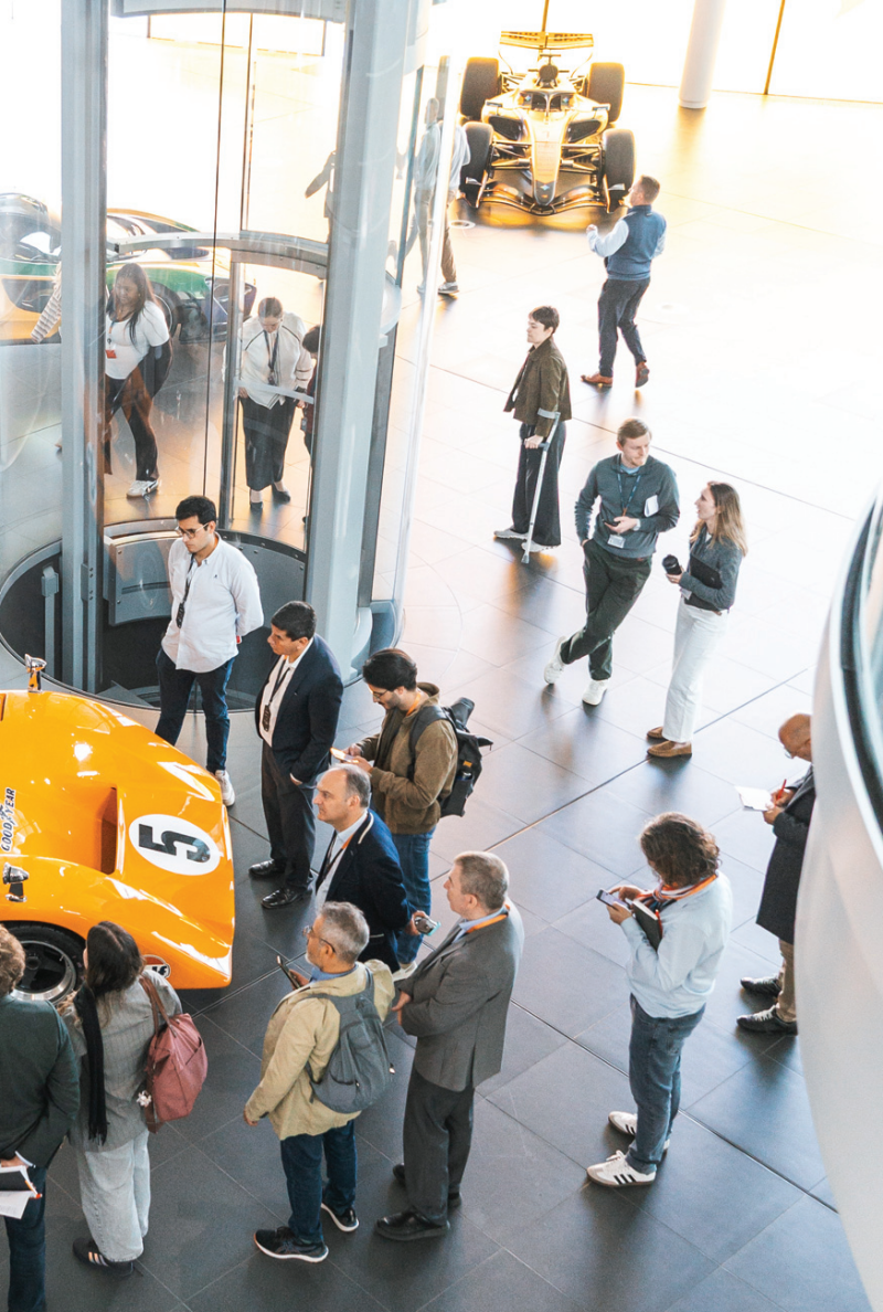 An overhead view of a corporate tour group looking at classic and modern McLaren racing cars displayed inside a bright, open facility showroom.