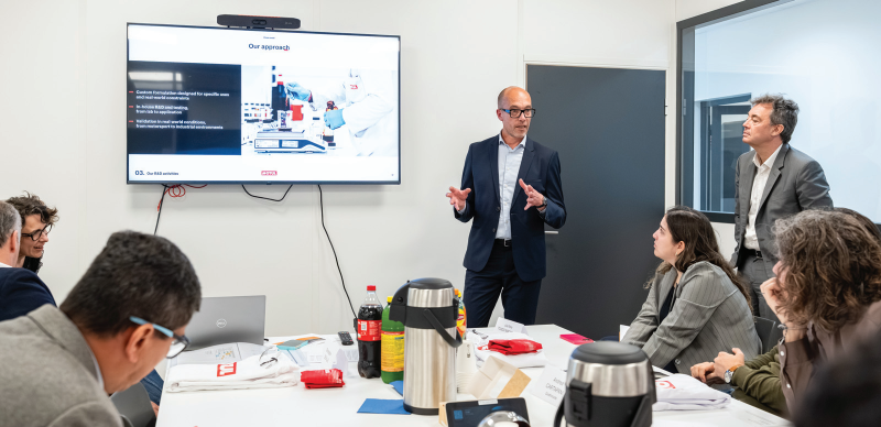 A business presentation inside a conference room with a speaker standing next to a large display screen showing research and development strategies to an attentive audience.