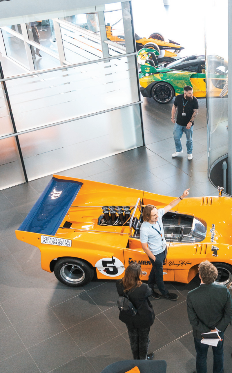 An overhead shot of a tour group looking at a vintage yellow McLaren race car displayed inside a modern corporate showroom.