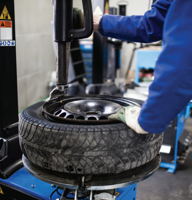 A mechanic using a tyre changer machine to service an automotive wheel and tyre assembly inside a garage workshop.