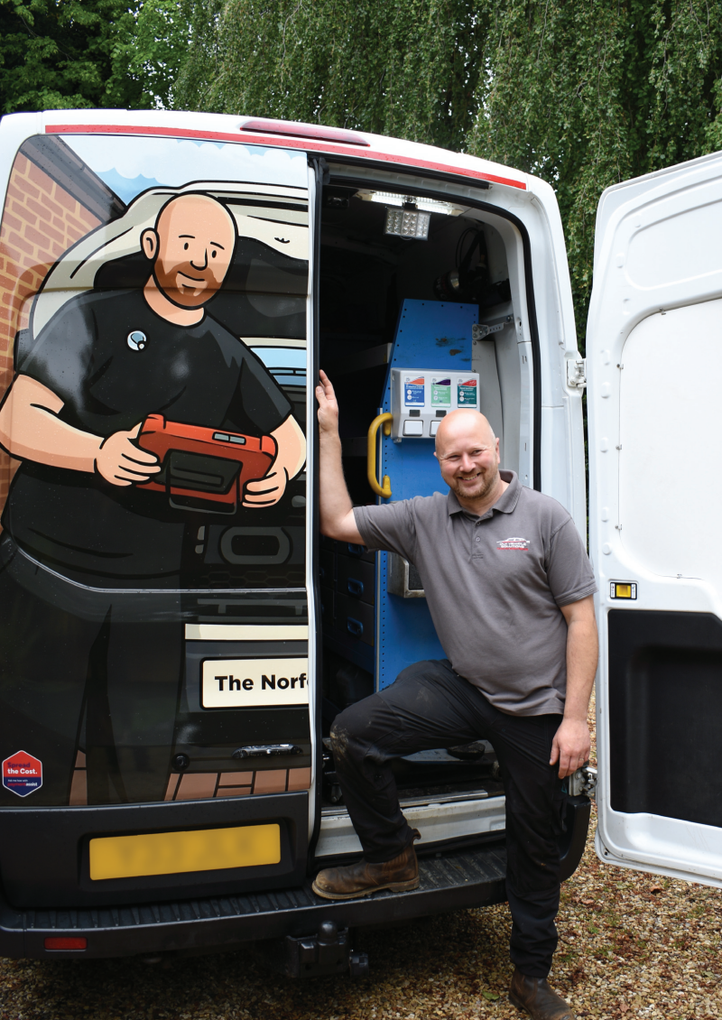 A mobile mechanic smiling next to his fully equipped service van featuring a custom cartoon graphic illustration on the rear door.
