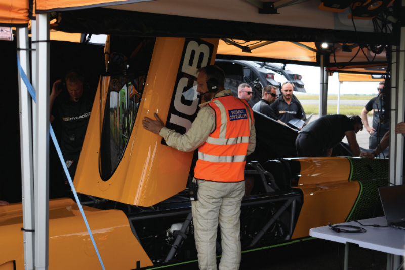 Support crew inspecting a custom yellow JCB high-speed vehicle under a canopy tent on an airfield.