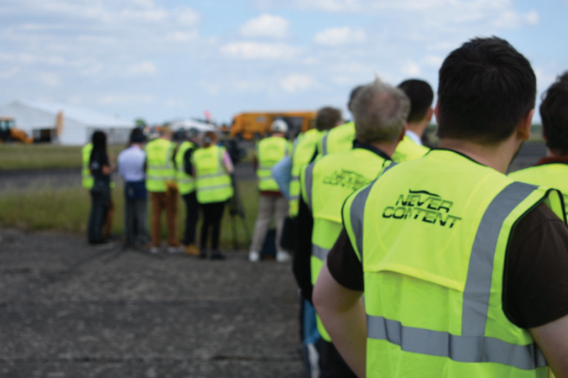 Spectators and crew members wearing high-visibility hi-vis vests lined up on an airfield tarmac watching an outdoor technical event.