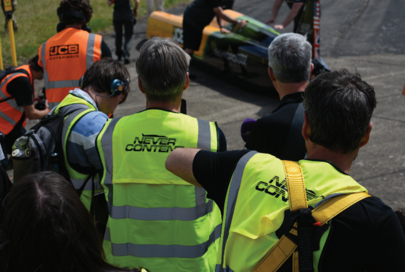 Overhead view of spectators and technical crew members in high-visibility vests watching a JCB high-speed vehicle on the tarmac.