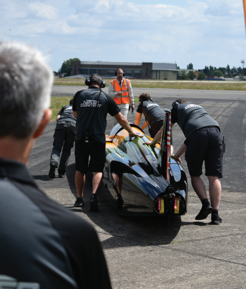 Support crew members pushing a black and yellow high-speed streamliner vehicle along an airfield runway.
