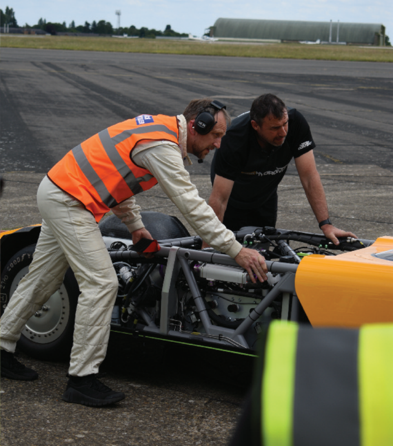 Two technicians inspecting the exposed tubular chassis and engine bay of a high-speed vehicle on an airfield tarmac.