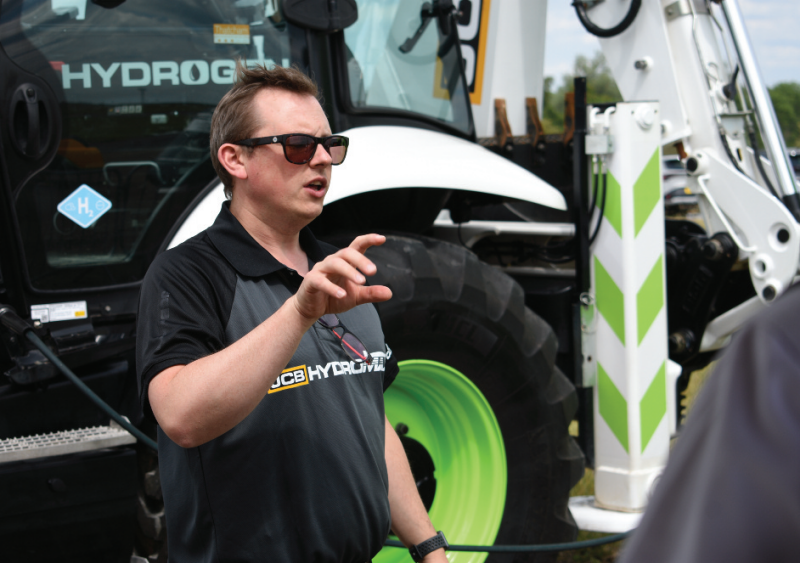 JCB Hydrogen engineer wearing sunglasses and a branded polo shirt speaking in front of a hydrogen-powered JCB backhoe loader.