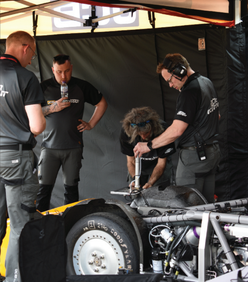 JCB pit crew members perform technical adjustments on an exposed high-speed streamliner chassis inside a canopy tent.