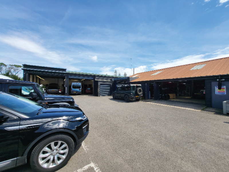 Wide-angle view of an automotive workshop facility forecourt with parked vehicles, open service bay doors, and a dark green 4x4.
