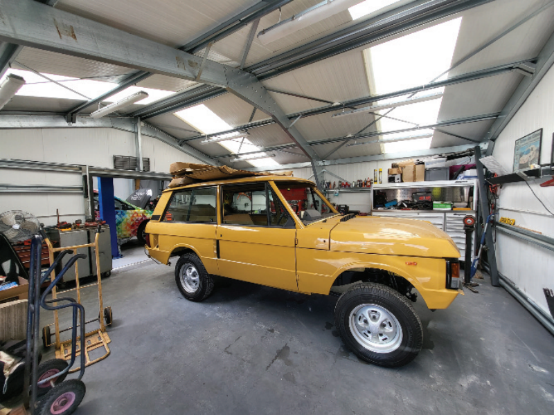 Classic yellow two-door Range Rover undergoing restoration inside a spacious automotive workshop with steel roof trusses and skylights.