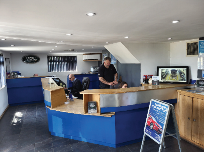 Automotive garage reception area featuring a curved blue service counter, staff members at workstations, and a prominent Land Rover wall emblem.