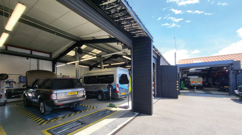 Automotive service bay with an SUV on test rollers alongside a passenger minibus near open bay doors leading to an outdoor courtyard.