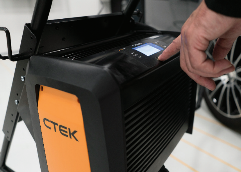 echnician pressing a button on the control panel of a CTEK professional battery charger mounted on a mobile workshop trolley.