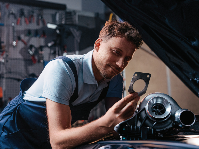 Automotive mechanic examining a metal exhaust flange gasket alongside a replacement turbocharger under an open vehicle bonnet.