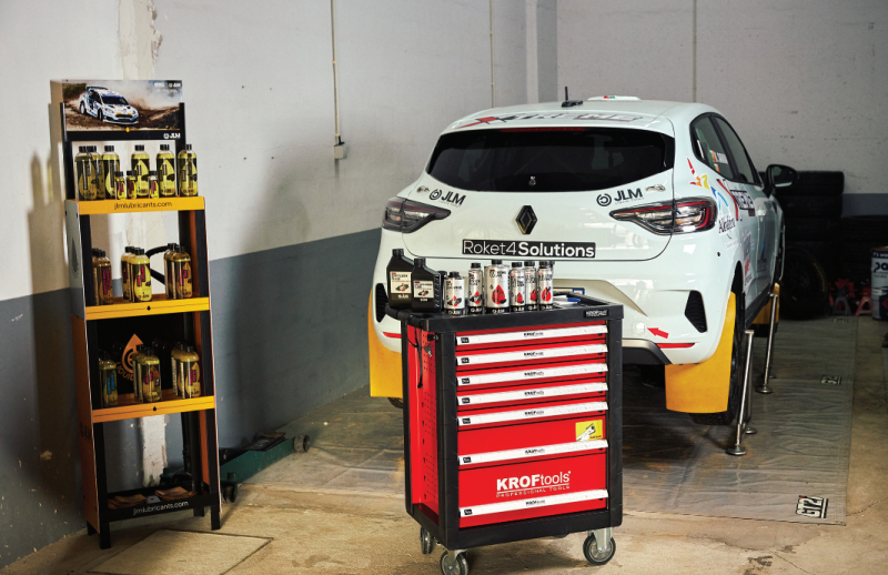White Renault rally car elevated on hydraulic alignment stands beside a Kroftools rolling tool cabinet and a JLM Lubricants retail display stand.
