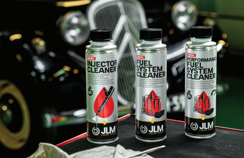 Three aluminum cans of JLM diesel additives—Diesel Injector Cleaner, Diesel Fuel System Cleaner, and Diesel Performance Fuel System Cleaner—arranged on a workshop surface in front of a classic car.