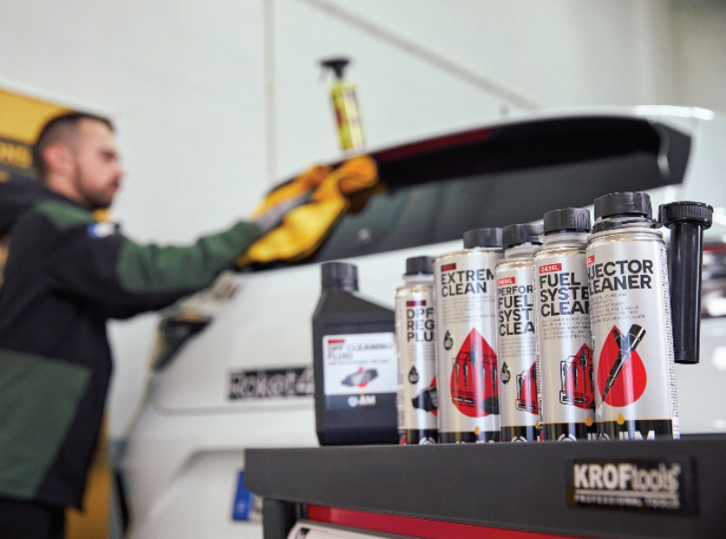 Lineup of JLM diesel maintenance additive bottles arranged on a Kroftools rolling cart while a technician wipes down a vehicle in the background.
