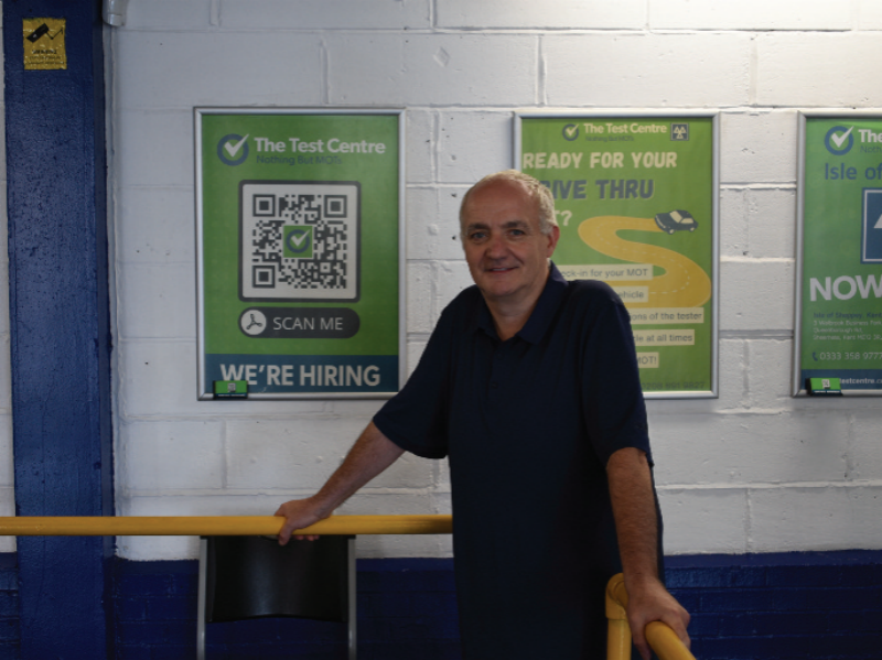 A man standing inside a workshop with white painted brick walls, leaning against a yellow safety rail next to promotional green posters for "The Test Centre", including a "We're Hiring" poster featuring a QR code.