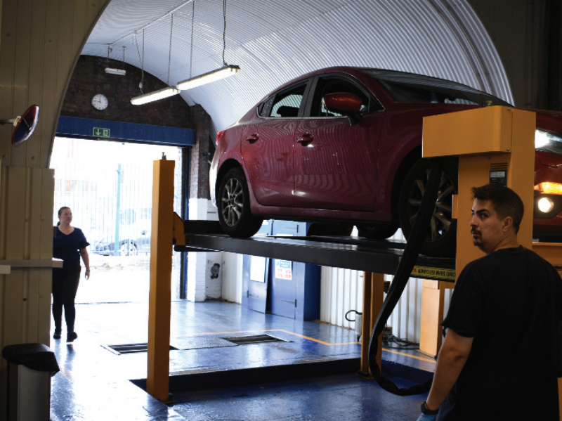 A red sedan lifted on a two-post hydraulic ramp inside a large arched garage workshop, with a technician standing near the lift pillar and a woman walking in the background near the open doorway.