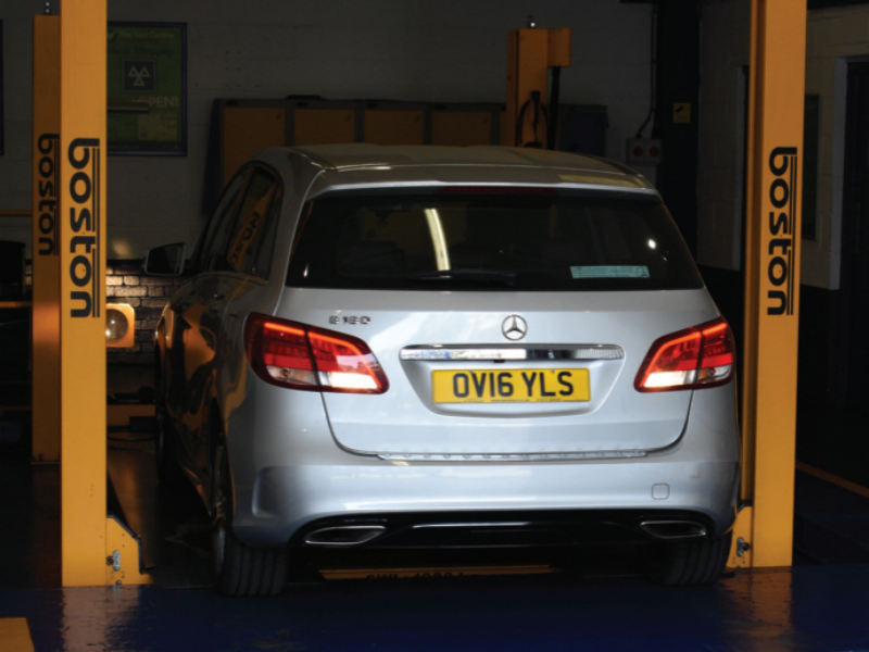 Rear view of a silver Mercedes-Benz B 180 parked between two yellow Boston two-post vehicle lift posts inside an automotive workshop, displaying registration plate OV16 YLS.