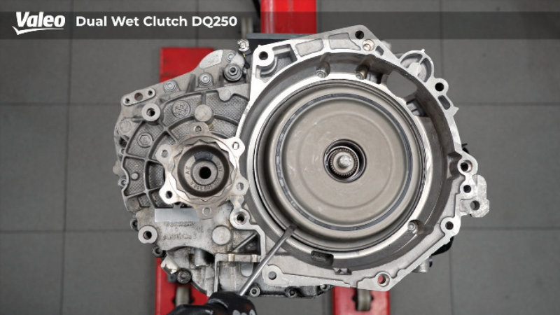 Top-down view of a Valeo Dual Wet Clutch DQ250 transmission assembly mounted on a workshop stand, showing the internal clutch housing, differential output flange, and a technician using a tool to check internal components against a grey tiled floor.