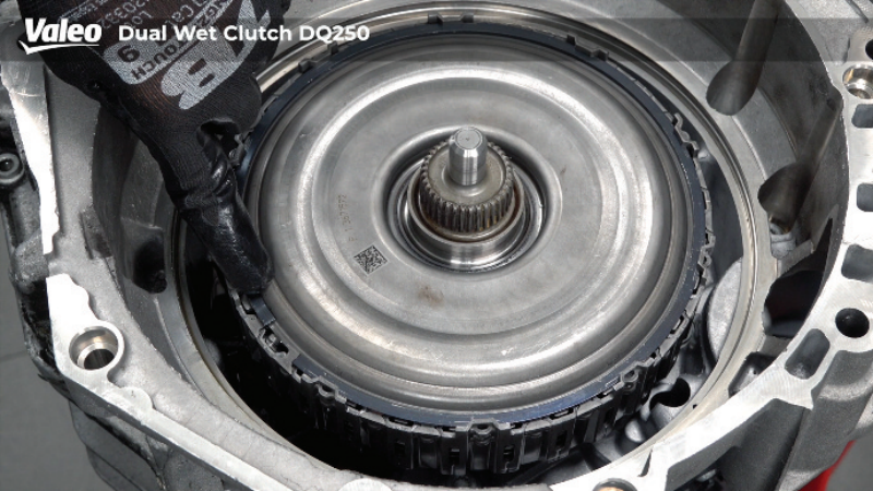 Close-up, top-down view of a Valeo Dual Wet Clutch DQ250 transmission assembly mounted on a workshop stand, with a technician wearing a black work glove pointing at the retaining clip ring on the internal clutch pack housing
