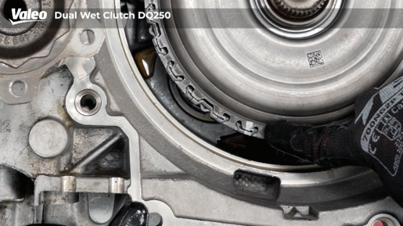 Close-up, top-down view of a Valeo Dual Wet Clutch DQ250 transmission assembly mounted on a workshop stand, featuring a technician wearing a black work glove pointing at the notched outer edge of the dual clutch assembly housing.