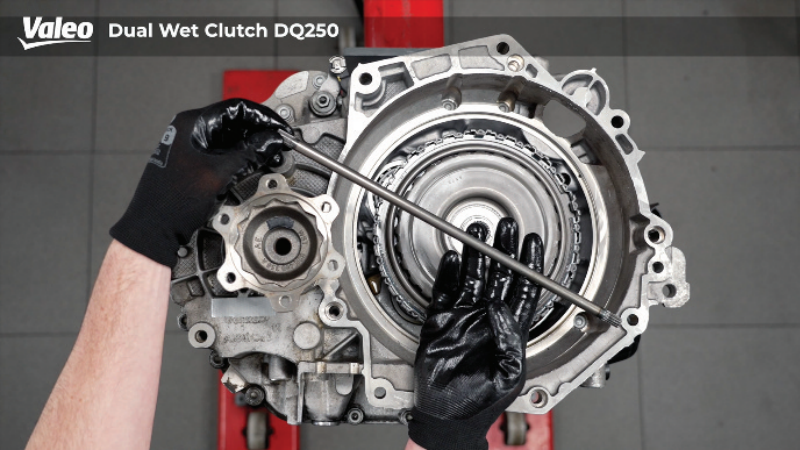 Top-down view of a Valeo Dual Wet Clutch DQ250 transmission assembly mounted on a workshop stand, featuring a technician wearing black work gloves holding a long alignment rod or tool horizontally across the internal clutch housing.
