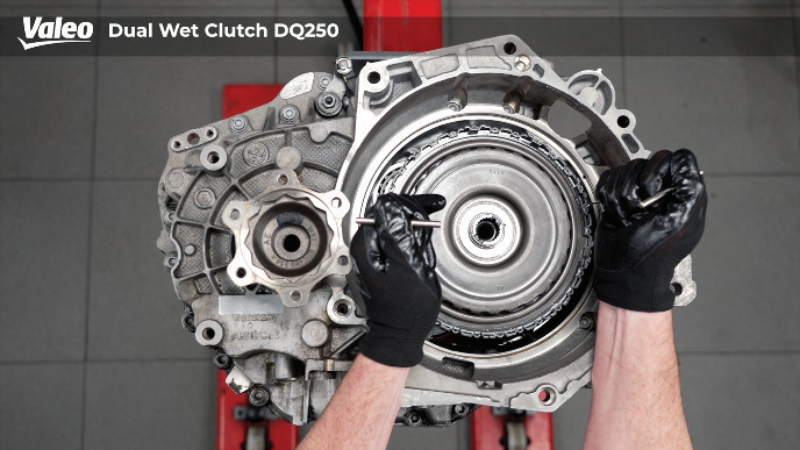 Top-down view of a Valeo Dual Wet Clutch DQ250 transmission assembly mounted on a workshop stand, with a technician wearing black work gloves using two alignment or extraction pins on opposing sides of the internal clutch pack housing.