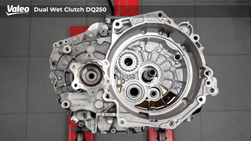 Top-down view of a Valeo Dual Wet Clutch DQ250 transmission assembly mounted on a workshop stand after the clutch pack has been fully removed, exposing the internal input shaft splines, roller bearings, and casing housing over a grey tiled floor.