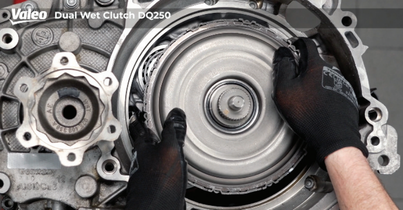 Top-down view of a Valeo Dual Wet Clutch DQ250 transmission assembly mounted on a workshop stand, featuring a technician wearing black work gloves carefully lowering or positioning the dual clutch assembly back into the transmission housing.