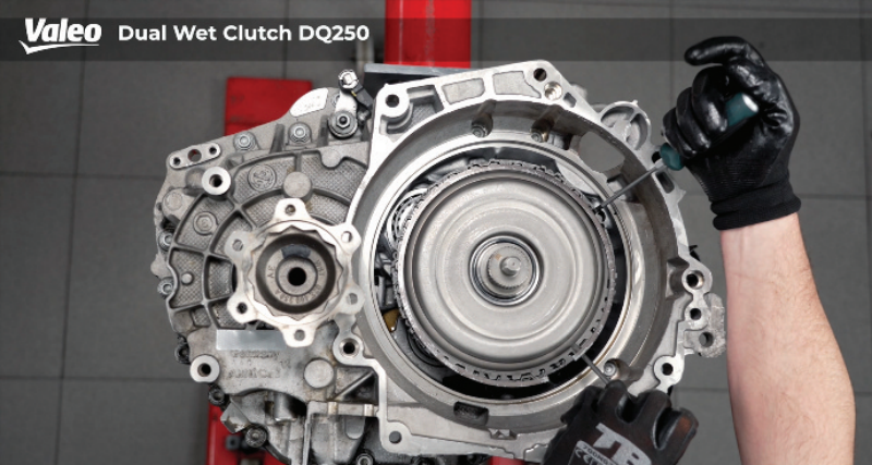 Top-down view of a Valeo Dual Wet Clutch DQ250 transmission assembly mounted on a workshop stand, featuring a technician wearing black work gloves using a tool to install or adjust the retaining mechanism around the clutch housing perimeter.
