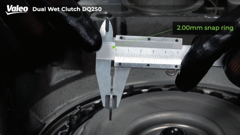Close-up view of a technician wearing black work gloves measuring a 2.00mm snap ring with a vernier caliper above the Valeo Dual Wet Clutch DQ250 assembly.