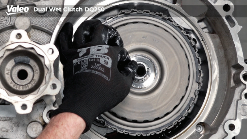 Top-down view of a Valeo Dual Wet Clutch DQ250 transmission assembly mounted on a workshop stand, featuring a technician wearing a branded black work glove gripping the central hub or extracting a component from the clutch pack housing.
