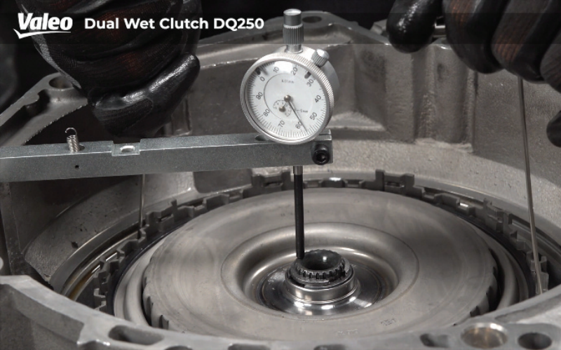 Close-up view of a technician wearing black work gloves measuring the axial play or clearance of the Valeo Dual Wet Clutch DQ250 assembly using a dial indicator gauge mounted on a bridge tool across the housing.
