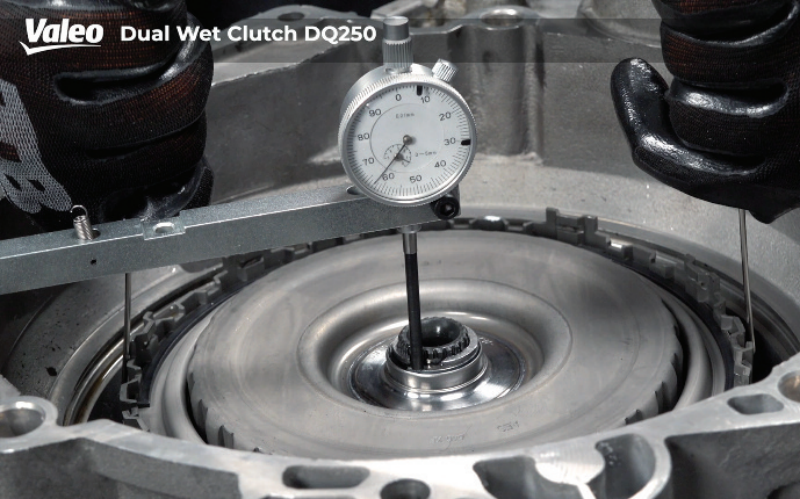 Close-up, wide-angle view of a technician wearing black work gloves measuring the axial play of the Valeo Dual Wet Clutch DQ250 assembly using a dial indicator gauge mounted on a bridge tool across the housing.