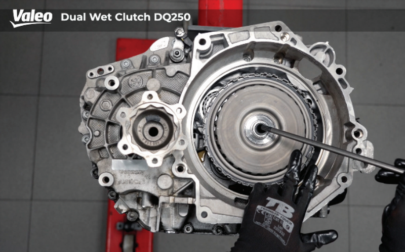 Top-down view of a Valeo Dual Wet Clutch DQ250 transmission assembly mounted on a workshop stand, featuring a technician wearing branded black work gloves using a long alignment tool or pry bar to adjust components within the housing perimeter.