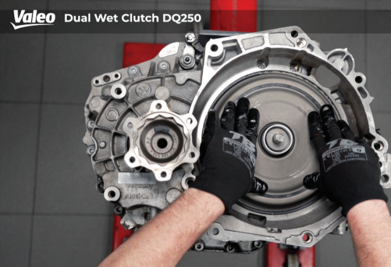 Top-down view of the Valeo Dual Wet Clutch DQ250 assembly mounted securely on a red workshop stand over a grey tiled floor, with a technician wearing branded black work gloves performing final checks or resting hands on the reinstalled clutch pack cover.